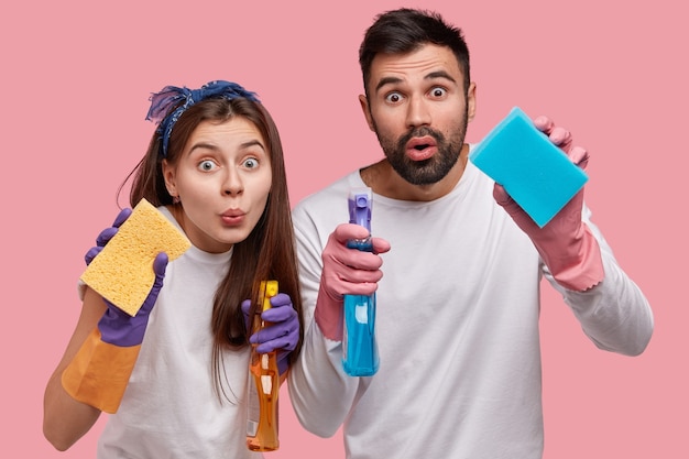 Horizontal shot of surprised handsome young guy and attractive woman holds sponges in front, carry detergents, clean windows at kitchen