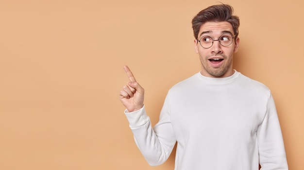 Horizontal shot of surprised handsome young man points at copy space shows awesome promotion or advertisement has amazed facial expression wears casual white jumper spectacles Wow look at this