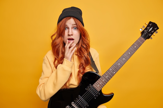 Horizontal shot of surprised redhead woman looks worried, wears sweatshirt and black hat poses with acoustic guitar 