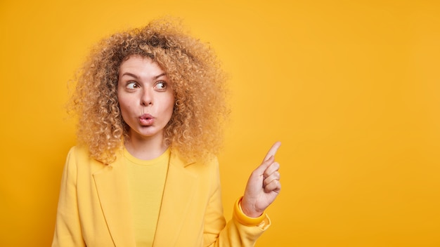 Free photo horizontal shot of surprised wondered woman with folded lips suggests to look away shows blank space for your advertisement dressed in formal clothes isolated over yellow wall. look there