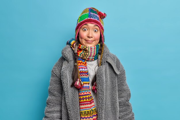 Horizontal shot of surprised young Asian woman with rosy cheeks wears knitted hat scarf around neck and warm fur coat dresses for cold weather isolated over blue wall