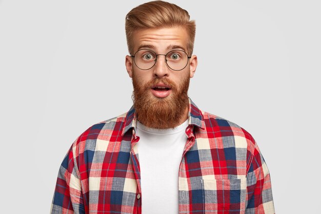 Horizontal shot of surprised young businessman has shocked expression, opens eyes widely, can`t beleive in failure, has thick ginger beard, wears checkered shirt, isolated over white wall