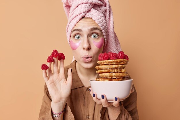 Horizontal shot of surprised young European woman stares wondered poses with yummy pancakes puts on raspberries on fingers applies hydrogel patches under eyes dressed in nightwear poses indoor