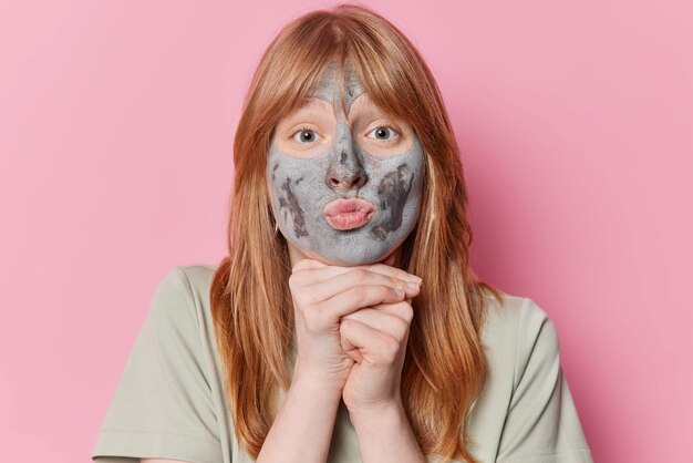 Horizontal shot of tender redhead girl keeps hands under chin has lips folded applies nourishing clay mask for rejuvenation and reducing fine lines isolated over pink background Beauty procedures