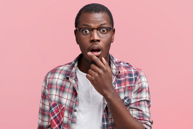 Horizontal shot of terrified male with shocked expression, keeps mouth opened and looks in bewilderment, wears glasses and casual t shirt, isolated over pink