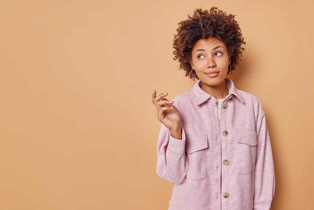 Horizontal shot of thoughtful curly haired woman keeps hand raised concentrated away and think about something with satisfied expression poses against beige background with blank copy space.