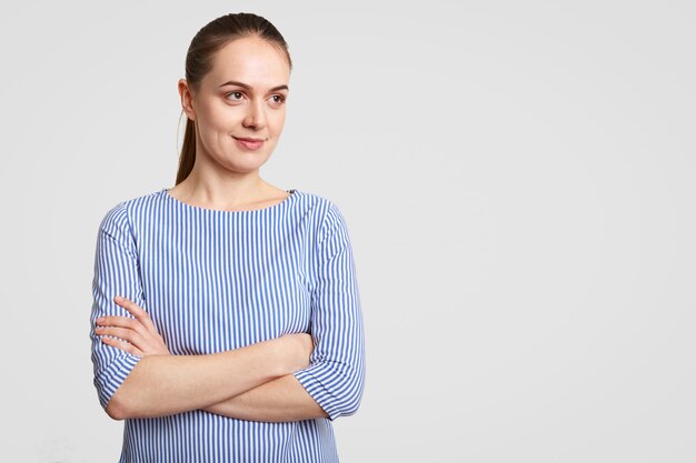 Horizontal shot of thoughtful European female with pony tail, keeps hands crossed, dressed in striped blouse