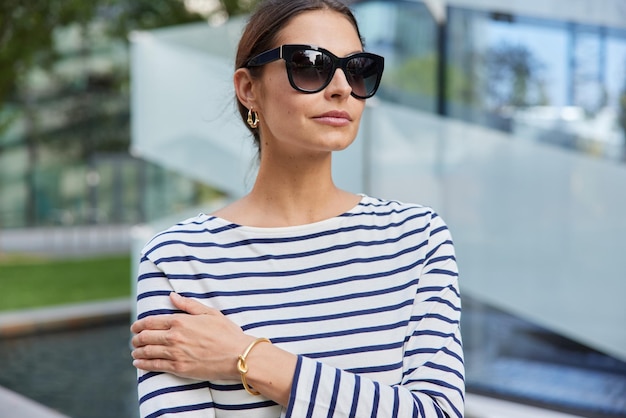 Horizontal shot of thoughtful European woman wears sunglasses casual striped jumper focused into distance walks outdoors poses at street against blurred background Street lifestyle concept