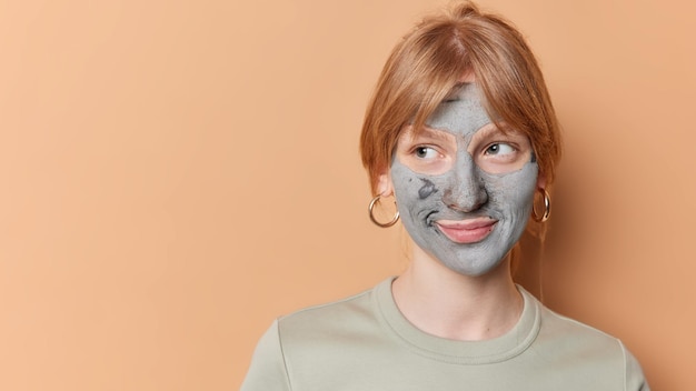 Horizontal shot of thoughtful ginger girl looks away undergoes beauty treatments applies clay mask for skin care dressed in casual t shirt poses against brown background blank space for your text