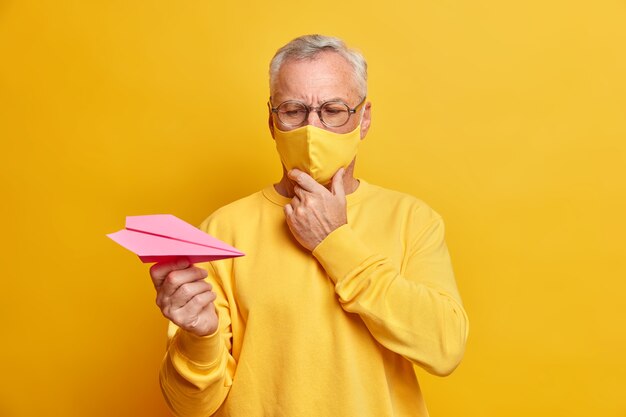 眼鏡をかけた思慮深い男性年金受給者の水平方向のショットは、紙飛行機を注意深く見ています。深刻な遠征があります。屋内での検疫ポーズ中に病気を克服する方法は保護マスクを着用します