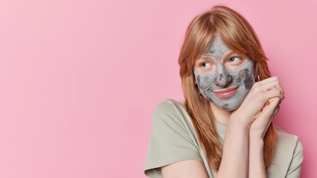 Horizontal shot of thoughtful red haired girl looks pensively away applies clay facial mask for skin treatments wears casual t shirt isolated over pink background blank copy space for your promo