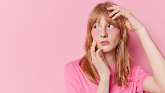 Horizontal shot of thoughtful redhead teenage girl keeps hand on face and head applies hydrogel patches under eyes undergoes beauty procedures has freckled skin isolated over pink background