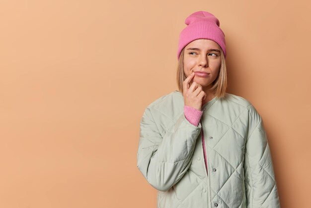 Horizontal shot of thoughtful woman considers something and looks away keeps finger near corner of lips wears pink hat and jacket isolated over brown background copy space on left feels hesitant