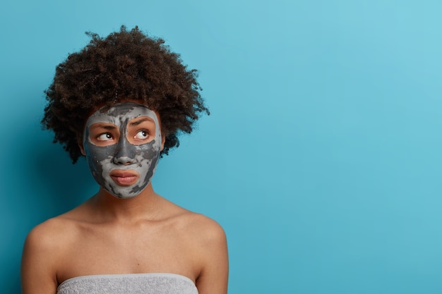 Horizontal shot of thoughtful woman with applied clay nourishing mask, looks aside, has bare shoulders, wrapped in bath towel, stands against blue wall with copy space. Beauty concept.