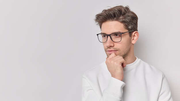 Horizontal shot of thoughtful young man holds chin looks attentively away ponders on something wears spectacles and jumper isolated over white background copy space for your promotional content