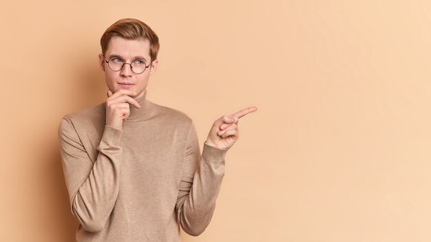 Horizontal shot of thoughtful young man holds chin thinks about suggested offer points away shows copy space for your advertising content wears round spectacles casual jumper 