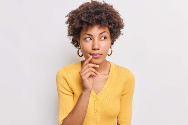 Horizontal shot of thoughtful young woman looks aside has dreamy expression interesting idea in mind wears casual yellow jumper isolated over white wall