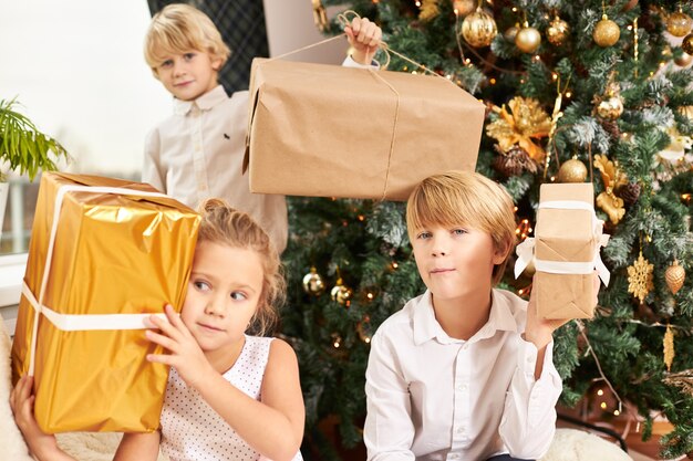 Горизонтальный снимок трех симпатичных братьев и сестер, сидящих на украшенной новогодней елке с коробками с рождественскими подарками, нетерпеливых и любопытных. Счастливое детство, радость и веселье
