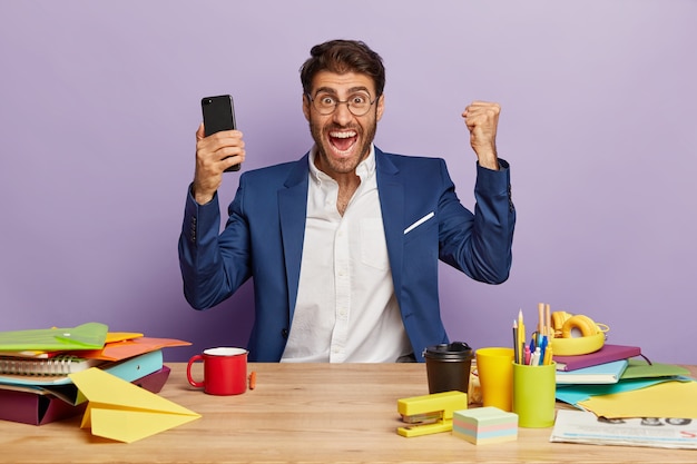 Horizontal shot of triumphing overjoyed businessman sitting at the office desk