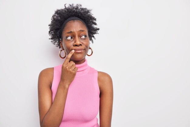 Horizontal shot of troubled thoughtful dark skinned woman keeps index finger near lips looksaway with worried pensive expression 