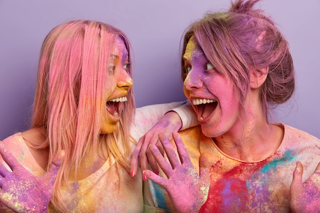 Horizontal shot of two happy women with colored hair, body and clothes, celebrate Holi Color festival, look happily at each other