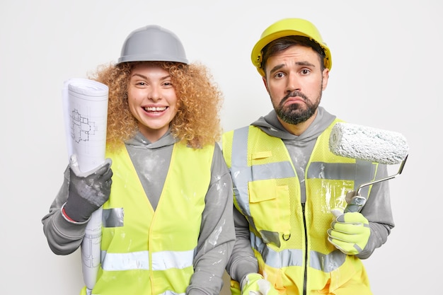 Horizontal shot of two professional maintenance workers paint walls of new building apartment hold painting roller use blueprint dressed in uniform remodels something 