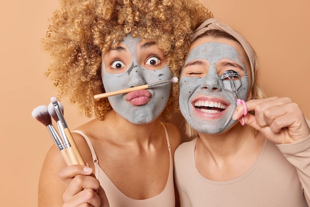 Free photo horizontal shot of two women undergo beauty procedures apply clay mask use cosmetic brushes and eyelashes curler isolated over brown background prepare for party skin care cosmetology concept