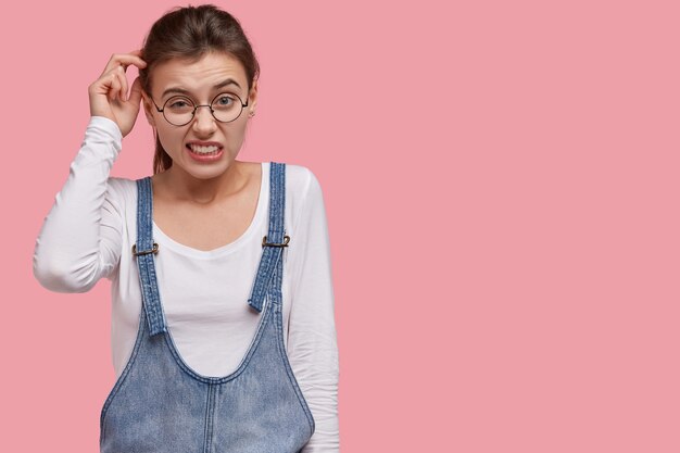 Horizontal shot of uncertain European woman scratches head with doubtful expression, tries to make decision, has displeased look, dressed in white jumper