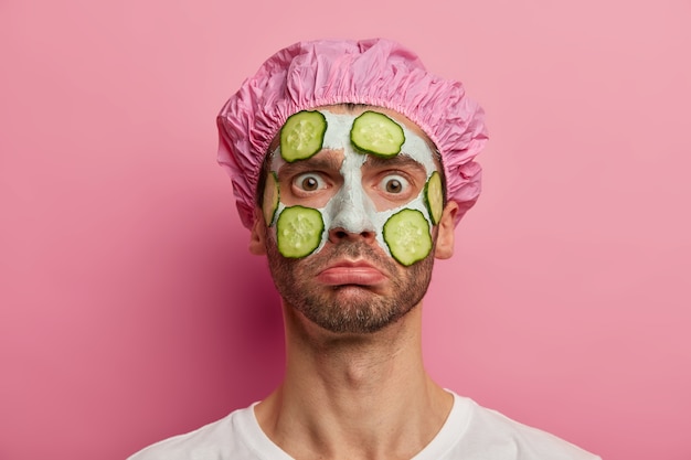 Free photo horizontal shot of unhappy man gets cosmetology treatments, refreshes skin with vegetable mask, stares at camera, wears bath hat