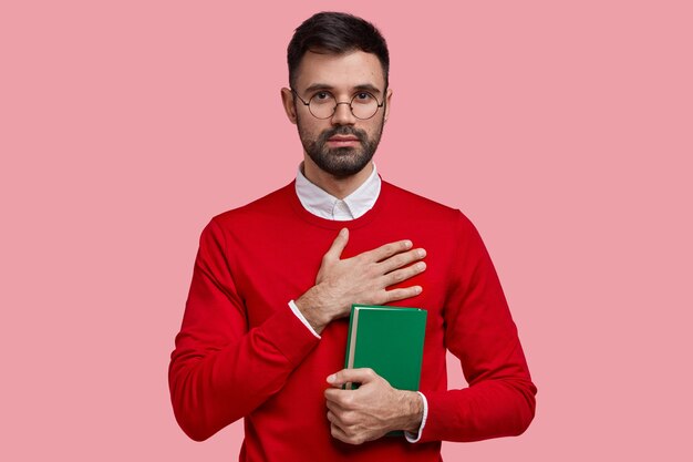 Horizontal shot of unshaven serious masculine college student promises to study hard, holds green textbook, wears red elegant sweater