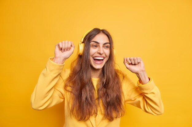 Горизонтальный снимок оптимистичной темноволосой женщины в наушниках, движущихся телом, наслаждается новым плейлистом, веселое выражение лица любит, когда мелодия носит желтый пиджак, широко улыбается, веселится