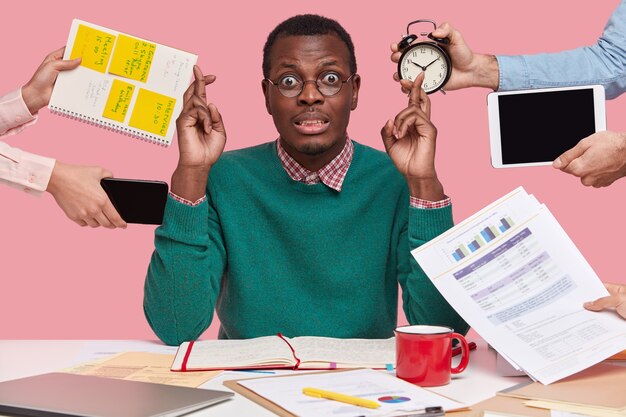 Horizontal shot of wishful dark skinned man crosses fingers, makes wish, dressed in green sweater, prepares for examination session, surrounded with alarm clock, touchpad