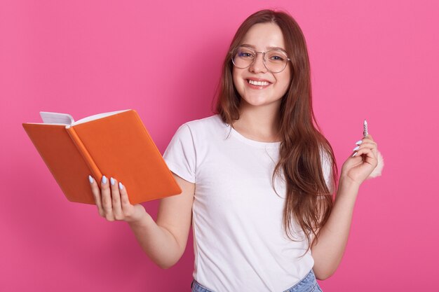 Horizontal shot of young attractive caucasian female in white casual clothes and spectacles, holding notebook and penil in hands