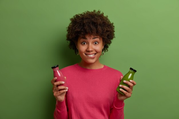 Horizontal shot of young dark skinned woman drinks green vegetable and pink fruit smoothie, bites lips, has healthy eating lifestyle, poses indoor in casual jumper. Weight loss, proper nutrition