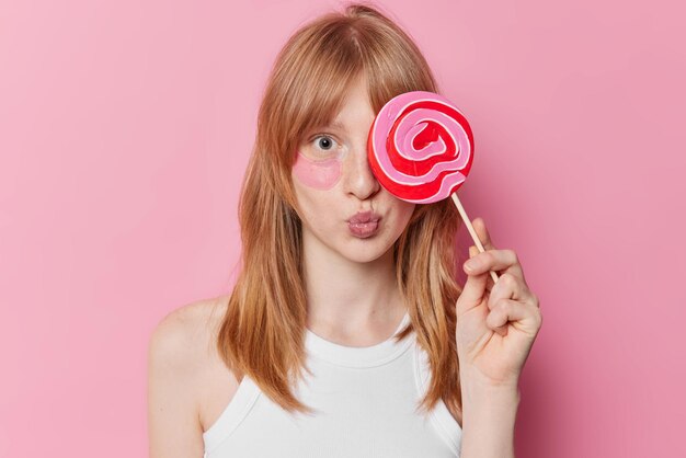 천연 진저 헤어 주근깨 피부를 가진 어린 소녀의 수평 사진은 눈 위에 카라멜 사탕을 덮고 분홍색 배경 뷰티 개념에서 격리된 붓기를 줄이기 위해 하이드로겔 패치를 적용합니다.