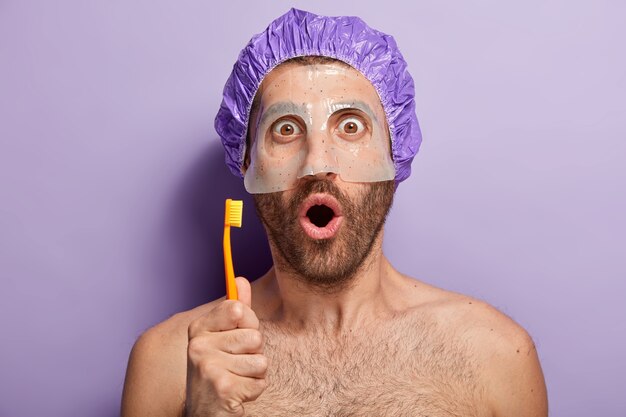 Horizontal shot of young man gets advice from professional cosmetologist, applies beauty mask for soft skin on face, going to brush teeth, holds toothbrush