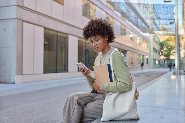 屋外に座っている若い女性の水平方向のショットは、スマートフォンのスクロールを真剣に見ていますカジュアルな服を着たソーシャルネットワークは、ストリートライフスタイルを歩いた後、街で布製のバッグの休符を運びます