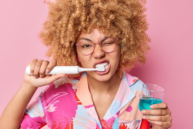 Horizontal shot of young woman with curly hair brushes teeth with electric toothbrush holds glass of fresh mouthwash takes care of dental hygiene isolated over pink background Daily routines