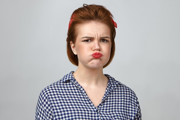 Horizontal studio portrait of irritated pretty girl in her twenties expressing dissatisfaction, blowing cheeks and pouting lips