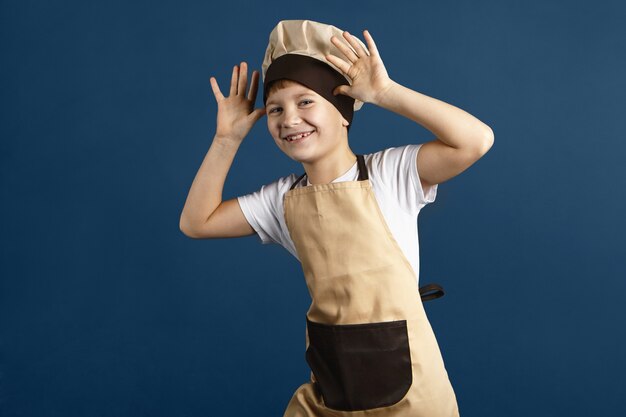 Горизонтальный студийный снимок забавного милого маленького мальчика, одетого в бежевую поварскую шляпу и фартук, смеющегося, держась за голову, корча рожи, дразня кого-то. Ребенок мужского пола с удовольствием. Концепция приготовления и питания