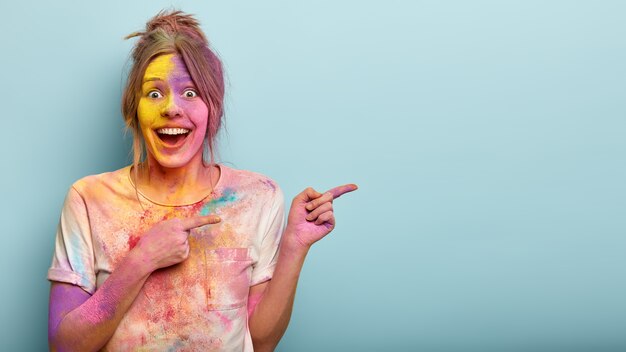 Horizontal studio shot of happy European woman points at side, covered with Holi colors