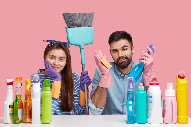 Horizontal view of attractive woman with dark hair, wears headband and protective gloves, sit closely to each other, surronded with cleaning agent