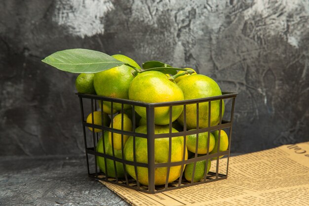 Horizontal view of black basket with fresh green tangerines and kumquats on newspapers on gray table