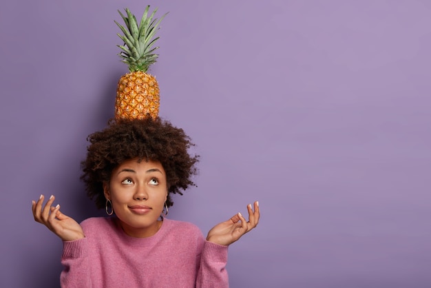 Free photo horizontal view of black curly woman tries to maintain equilibrium, holds fresh pineapple on head, spreads palms, looks above