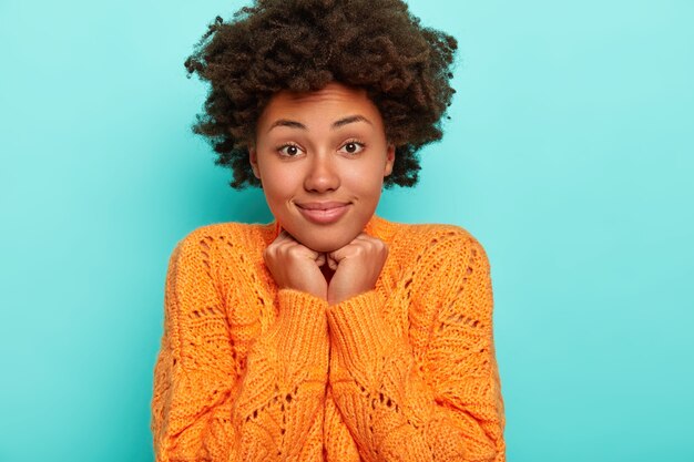 Horizontal view of cheerful curious surprised African American lady with dark bushy hair, touches chin with both hands, dressed in vivid orange sweater, isolated on blue wall