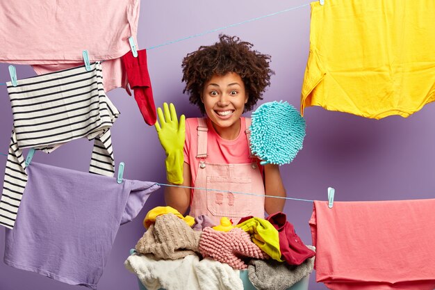 Horizontal view of cheerful curly haired woman busy doing work about house, wears protective rubber gloves, holds mop for cleaning, hangs laundry after washing, happy almost finishing working