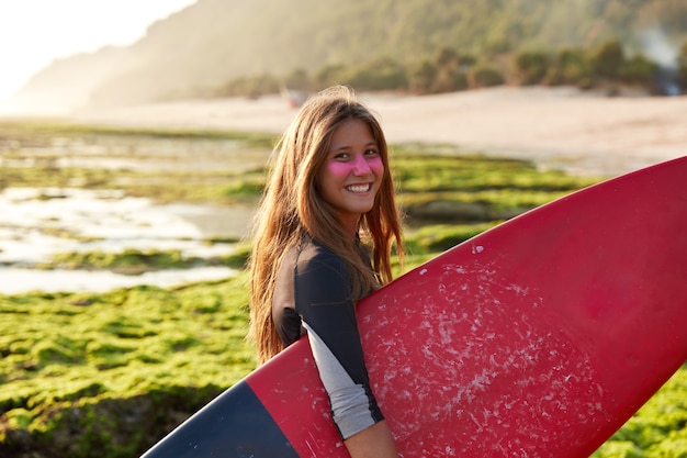 Horizontal view of cheerful European surfer surfes being in high spirit, carries board