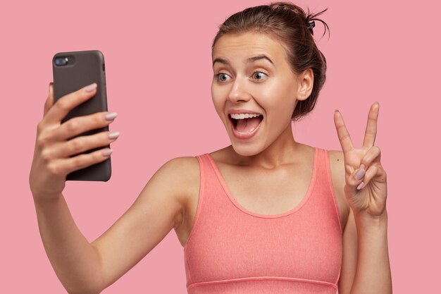 Horizontal view of cheerful European woman makes peace gesture of cell phone, dressed in pink vest, being in good mood