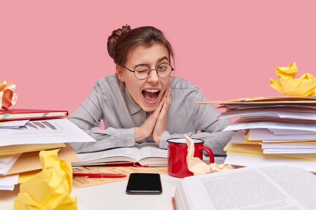 Free photo horizontal view of cheerful schoolgirl blinks eyes, wears round spectacles, keeps hands together, has fun during studying process