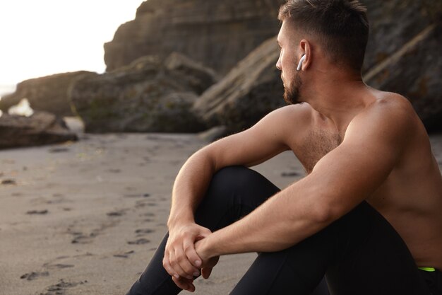 Горизонтальный вид созерцающего спортсмена, сидящего на песке, сфокусированного вдаль
